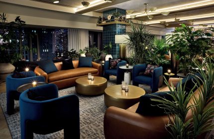 Viceroy Chicago, a gay-friendly hotel. Stylish lounge area with leather sofas, blue armchairs, and lush greenery. Nighttime city view.