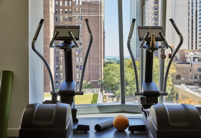 Viceroy Chicago gay-friendly hotel gym with elliptical machines, yoga mat, and ball. City view visible through the window.