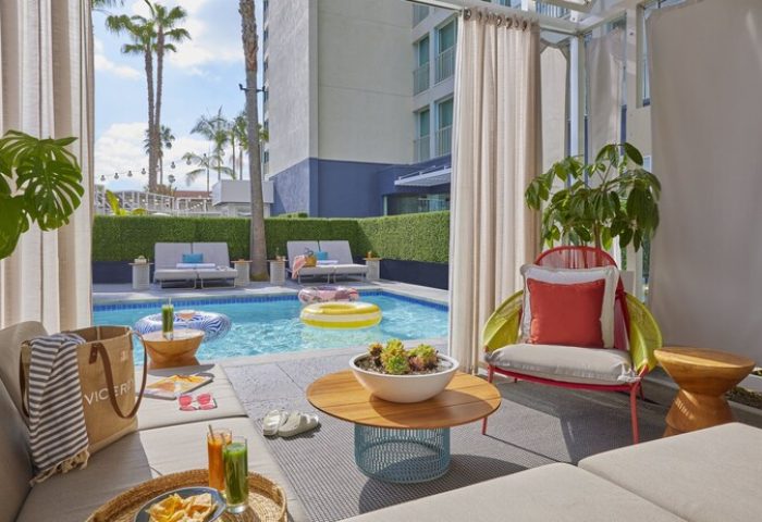 Viceroy Santa Monica gay-friendly hotel poolside cabana view. Relaxing by the pool with drinks, snacks, and stylish decor. Palm trees in the background.