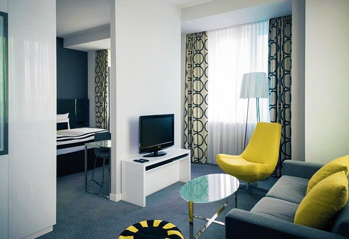 Vienna House Andel’s Berlin room. A gay-friendly, pet-friendly hotel. Modern design with yellow chair, sofa, and TV. Bedroom view.