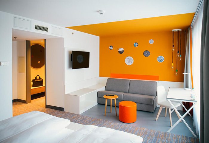 Vienna House Easy Berlin gay-friendly hotel room. Modern design, orange accents, sofa, desk, and TV. Perfect for gay travelers in Berlin.