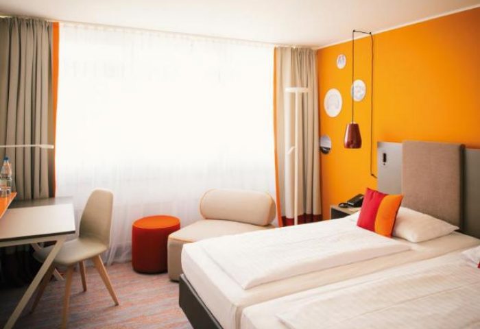 Vienna House Easy Berlin room. Gay-friendly hotel with twin beds, desk, chair, orange wall, and modern decor. A comfortable stay in Berlin.