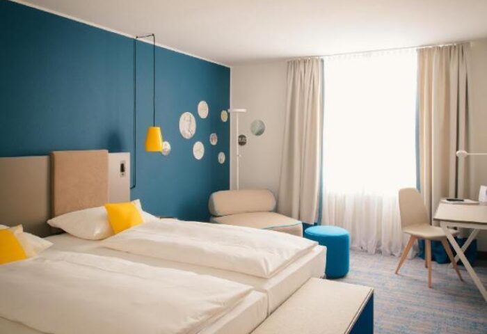 Bedroom at Vienna House Easy Berlin, a gay-friendly hotel. Blue wall, double bed, desk, chair, and decor. Modern, clean design.
