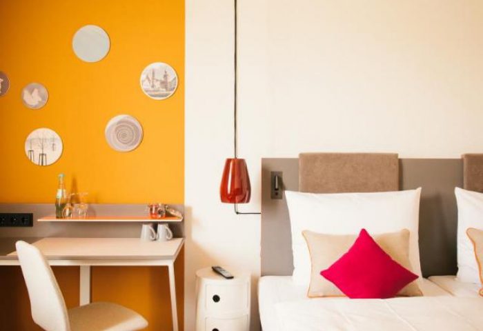 Vienna House Easy Berlin gay-friendly hotel room. Desk, bed with pillows, and orange wall. A comfortable stay for gay travelers in Berlin.