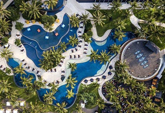 Aerial view of the W Bali Seminyak, a gay-friendly hotel. Features pools, palm trees, lounge chairs, and a round building.
