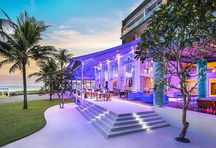 W Bali Seminyak gay-friendly hotel outdoor restaurant. Evening view with purple lighting, palm trees, and ocean. Gay travel advice.
