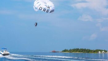 Parasailing at W Maldives, a gay-friendly hotel. Boat pulling a person with a 'Dream' parachute over the ocean. Tropical island view.