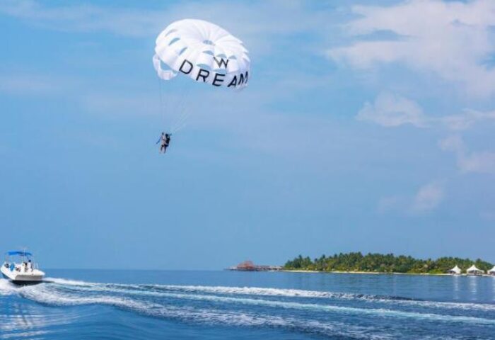 Parasailing at W Maldives, a gay-friendly hotel. Boat pulling a person with a 'Dream' parachute over the ocean. Tropical island view.
