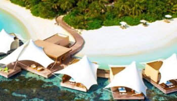 W Maldives gay-friendly hotel aerial view. Overwater bungalows with sunshades, white sand beach, and lush tropical vegetation. Luxury gay travel.