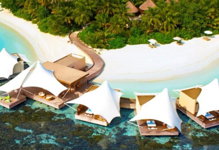 W Maldives gay-friendly hotel aerial view. Overwater bungalows with sunshades, white sand beach, and lush tropical vegetation. Luxury gay travel.