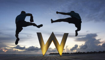 Two men jumping over the W Maldives logo at sunset. Gay-friendly hotel with overwater bungalows. A perfect gay travel destination.