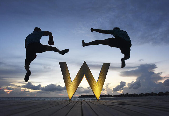 Two men jumping over the W Maldives logo at sunset. Gay-friendly hotel with overwater bungalows. A perfect gay travel destination.