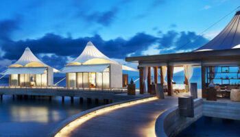 W Maldives gay-friendly hotel. Overwater bungalows with tented roofs on a wooden walkway at dusk. Luxury travel destination for gay men.