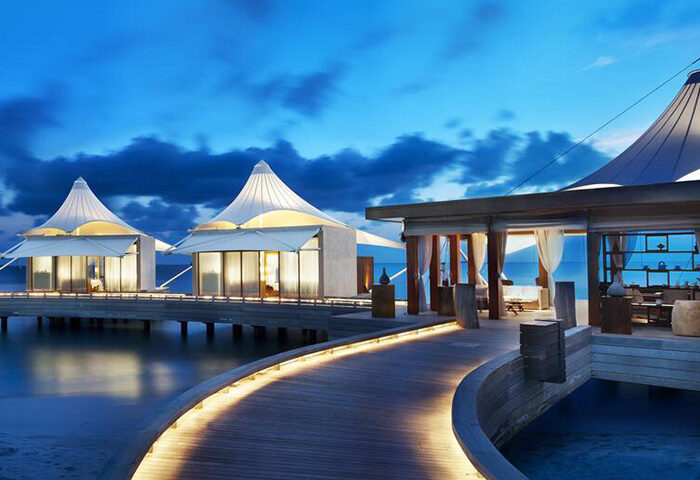 W Maldives gay-friendly hotel. Overwater bungalows with tented roofs on a wooden walkway at dusk. Luxury travel destination for gay men.