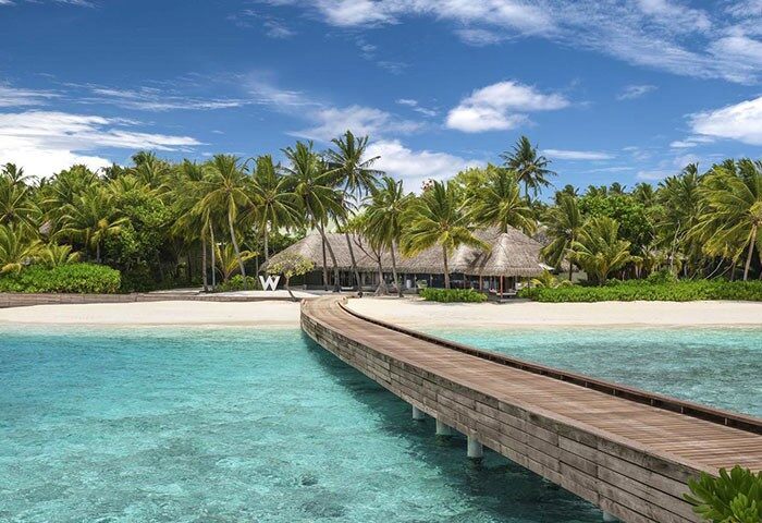 Wooden pier at W Maldives, a gay-friendly hotel. Tropical island with palm trees, white sand beach, and turquoise water. Perfect for LGBTQ+ travel.
