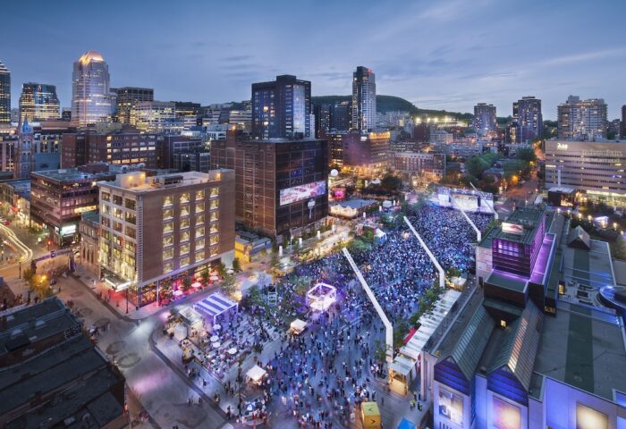 Night view of W Montreal, a gay-friendly hotel, overlooking a crowded outdoor event in the city. Montreal's skyline is visible.