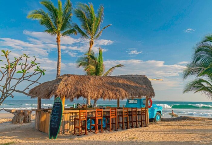 Beach bar at W Punta de Mita, a gay-friendly hotel. Thatched roof, blue truck, and surfboard on the sand. Palm trees and ocean backdrop.