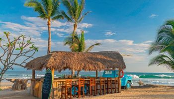 Beach bar at W Punta de Mita, a gay-friendly hotel. Thatched roof, blue truck, and surfboard on the sand. Palm trees and ocean backdrop.