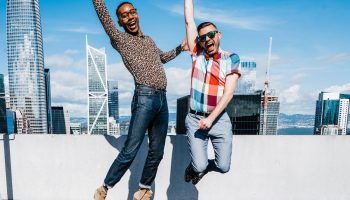 Two gay men jumping for joy at W San Francisco hotel. Celebrating LGBTQ travel in a gay-friendly hotel with city skyline view.