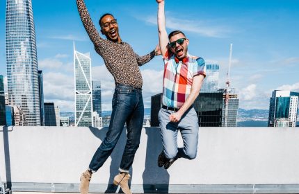 Two gay men jumping for joy at W San Francisco hotel. Celebrating LGBTQ travel in a gay-friendly hotel with city skyline view.