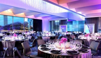 W San Francisco hotel ballroom setup for a gay wedding. Tables with flowers, glasses, and black napkins fill the room. Purple lighting.