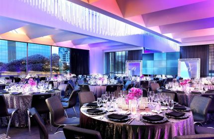 W San Francisco hotel ballroom setup for a gay wedding. Tables with flowers, glasses, and black napkins fill the room. Purple lighting.