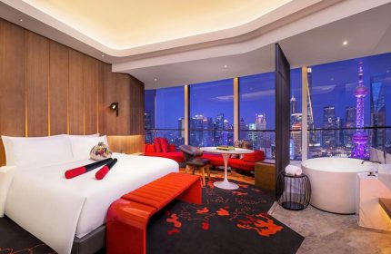 W Shanghai The Bund hotel room interior at night. Gay-friendly hotel with city view, bed, red accents, and bathtub. Shanghai skyline.