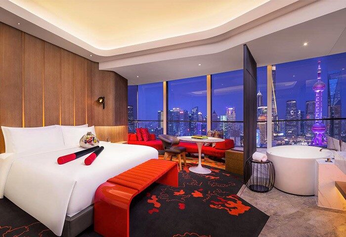 W Shanghai The Bund hotel room interior at night. Gay-friendly hotel with city view, bed, red accents, and bathtub. Shanghai skyline.
