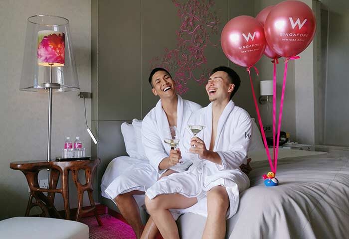 Gay couple at W Singapore - Sentosa Cove, a gay-friendly hotel. They're laughing, wearing robes, and holding cocktails with balloons.