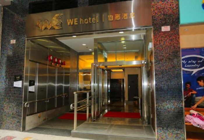 Entrance to WE Hotel, a gay-friendly hotel. Modern design with stainless steel and glass doors. Red carpets welcome guests. Urban setting.