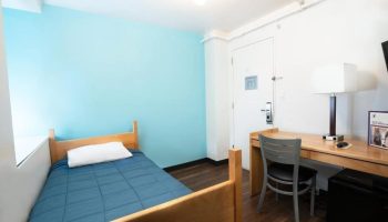 West Side YMCA Hostel room. Gay-friendly hotel with single bed, desk, chair, and blue accent wall. Budget-friendly lodging for gay travelers.