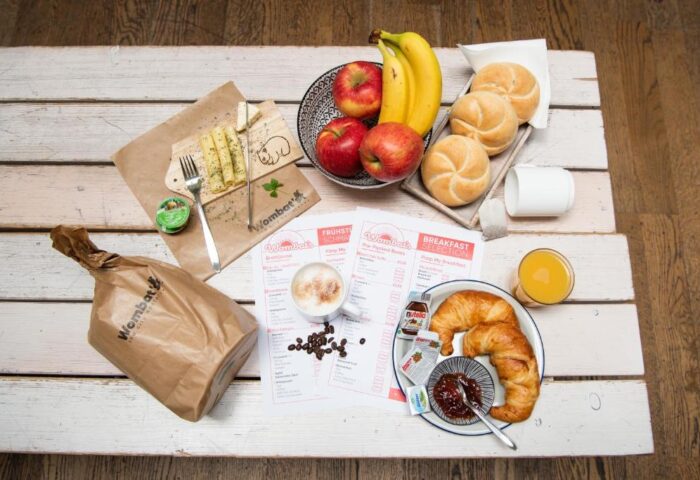 Breakfast at Wombats City Hostel London, a gay-friendly hotel. Fruit, croissants, rolls, cheese, and coffee on a white wooden table.