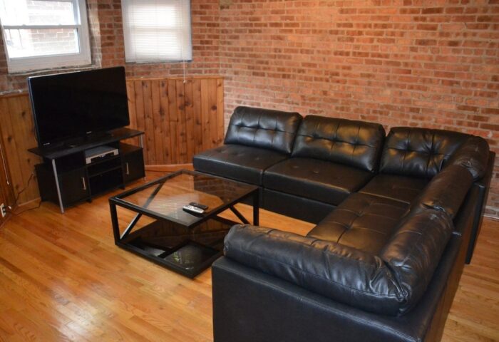 Wrigley Hostel Adults Only gay-friendly hotel room. Black leather sectional sofa, glass coffee table, TV. Brick wall, wood paneling, hardwood floor.