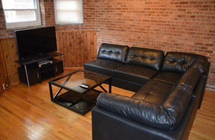Wrigley Hostel Adults Only gay-friendly hotel room. Black leather sectional sofa, glass coffee table, TV. Brick wall, wood paneling, hardwood floor.
