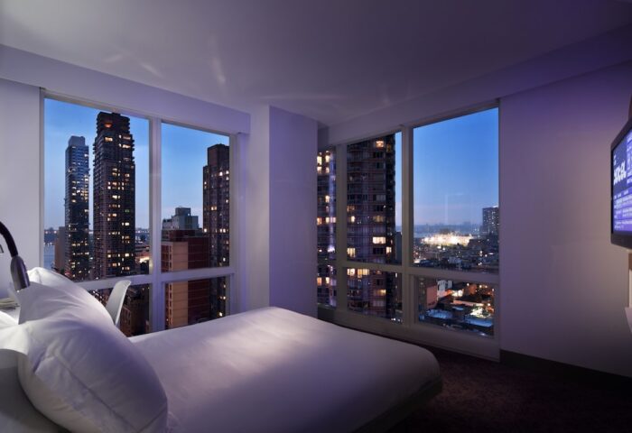 YOTEL New York Times Square gay-friendly hotel room at night. City skyline view from bed. Modern design for gay travelers.