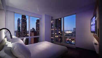 YOTEL New York Times Square gay-friendly hotel room at night. City skyline view from bed. Modern design for gay travelers.