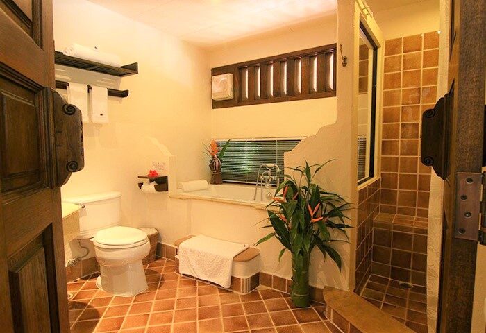 Bathroom at Yaang Come Village Hotel, a gay-friendly hotel. Features toilet, tub, shower, brown tile floor, and tropical plants.