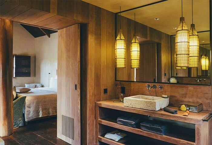 Zannier Hotels Phum Baitang bathroom. Stone sink, wooden shelves with towels, and woven pendant lights. Gay-friendly hotel room interior.