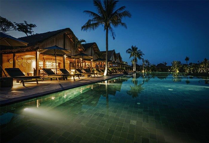 Zannier Hotels Phum Baitang pool at night. Gay-friendly hotel with luxury villas, palm trees, and deck chairs. Ideal for gay men's travel.
