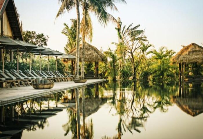 Gay-Friendly Zannier Hotels Phum Baitang pool view. Tropical resort with palm trees, lounge chairs, and thatched-roof pavilions. Serene reflection.