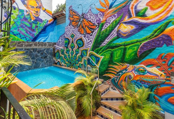 ZonaZ Boutique Hotel's gay-friendly pool area. Colorful mural with butterfly, lizard, and portrait. Tropical plants add to the vibrant scene.