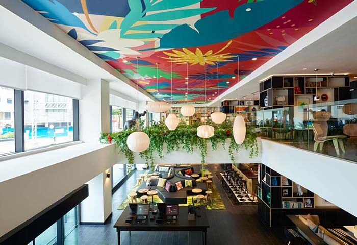 citizenM Kuala Lumpur Bukit Bintang interior. Gay-friendly hotel with colorful ceiling art, hanging lights, and lush greenery. Modern design.