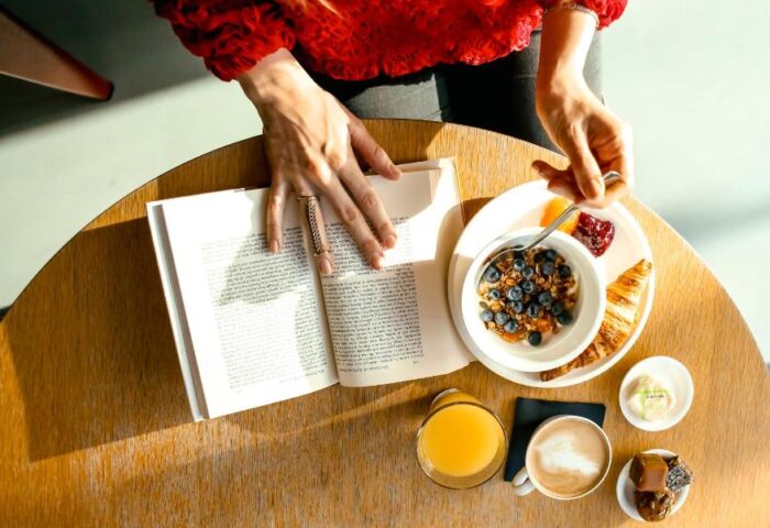 Breakfast at citizenM London Shoreditch, a gay-friendly hotel. Woman reads a book while eating granola, croissant, and drinking juice and coffee.