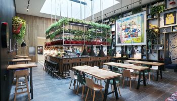 citizenM Los Angeles Downtown gay-friendly hotel bar area with tables, chairs, and greenery. Modern design with skylight. Great for gay travel.