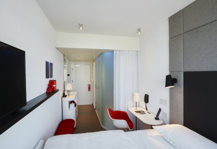 citizenM New York Bowery hotel room. Modern design with white walls, red accents, desk, and bed. Gay-friendly hotel for travelers.