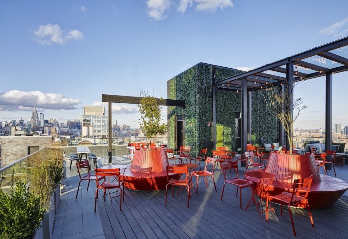 citizenM New York Bowery gay-friendly hotel rooftop. Red chairs and tables on a wooden deck with NYC skyline in the background. Outdoor seating.