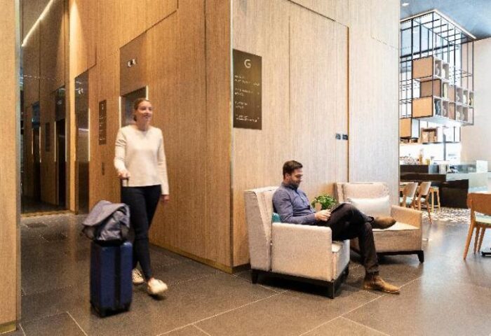 Ibis Melbourne Central lobby with guests. A woman arrives with luggage, while a man relaxes in a chair. Modern, gay-friendly hotel design.