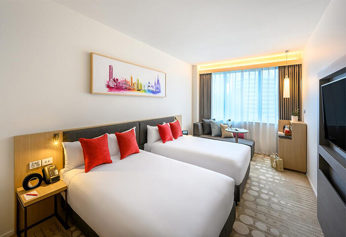 Ibis Melbourne Central hotel room with two beds, red pillows, and a couch by the window. Gay-friendly hotel with modern decor.