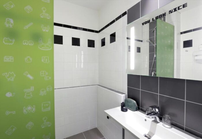 Ibis Styles Budapest Center bathroom. Modern design with white & black tiles, green accent wall with game icons. Mirror says You Look Nice.