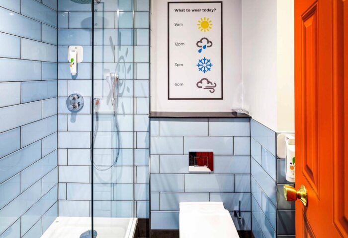 Ibis Styles Manchester Portland gay-friendly hotel bathroom. Shower, toilet, and What to wear today? weather forecast. Orange door.